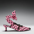 A pink pointed-toe heeled mule with black polka dots, featuring a matching polka-dot ankle strap tied in a large bow, photographed on a dark gray gradient studio background.