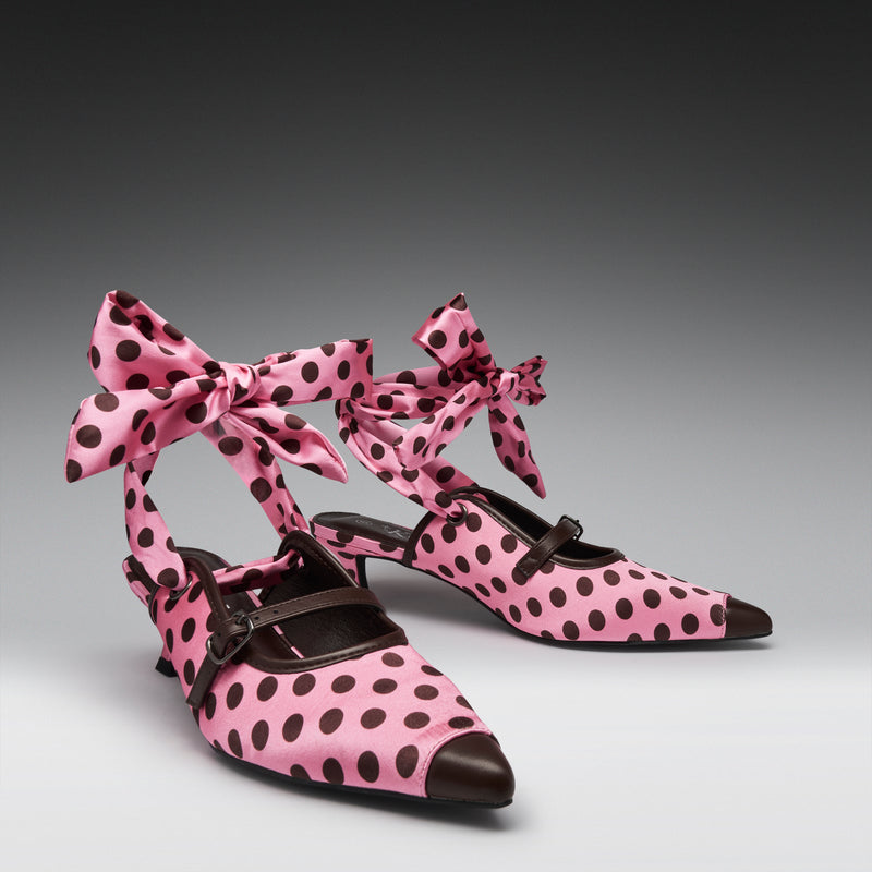 Pink pointed-toe Mary Jane-style heels with dark brown polka dots, bow ankle straps, and dark brown leather trim, photographed on a gray gradient studio background.
