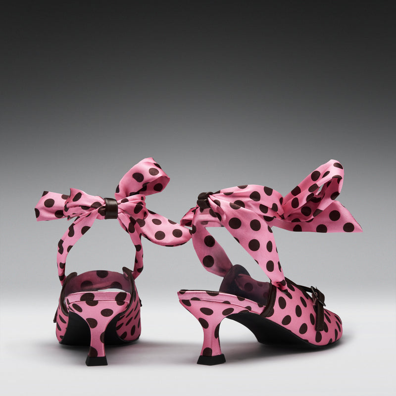 A pair of pink satin high-heeled sandals with black polka dots and large matching bow straps, shown from the back on a dark-to-light gray gradient studio background.