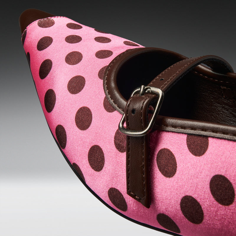A close-up of a pink pointed-toe shoe with dark brown polka dots, a dark brown strap, and a silver buckle.