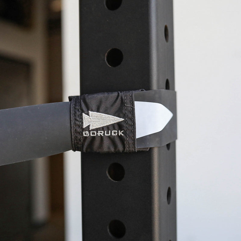 A Recovery Floss Band labeled "GORUCK" secures a bar to a rack with evenly spaced holes, making it ideal for muscle compression and mobility improvement techniques.