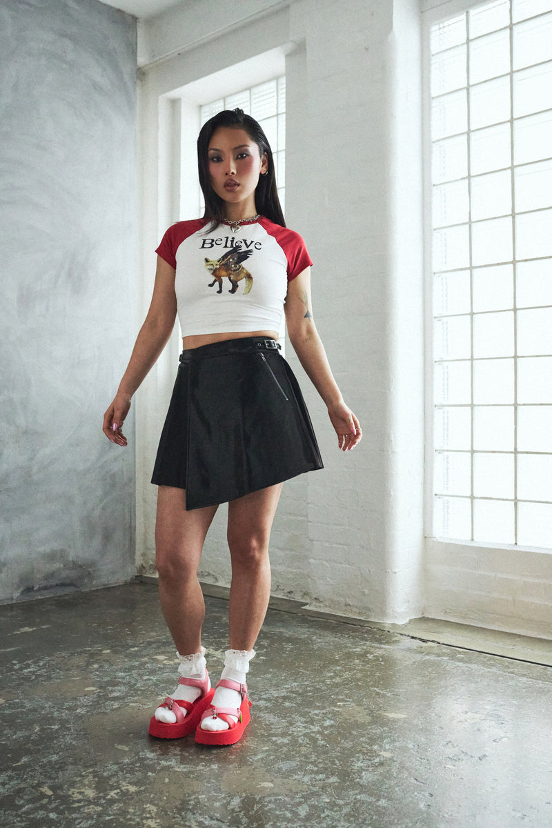 Woman wearing a white and red crop top with a graphic design, black skirt, and red sandals in a room with large windows.
