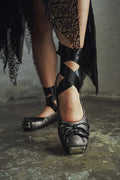 Black high-heeled shoes with criss-cross straps worn by a person on a textured floor.