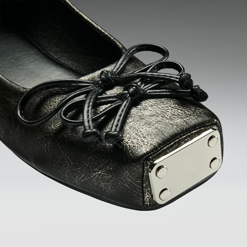 Black leather shoe with a decorative bow and metallic heel tab on a gray background