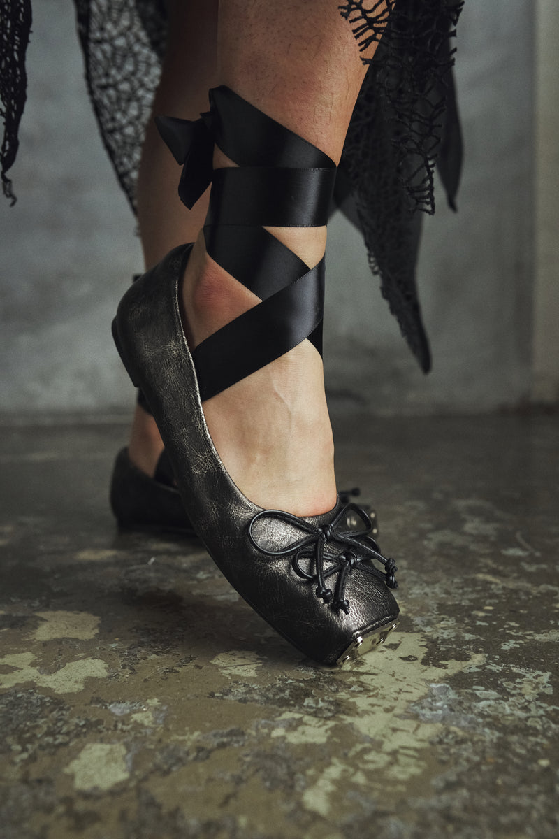 Black ballet flats with criss-cross straps worn by a person on a textured floor.