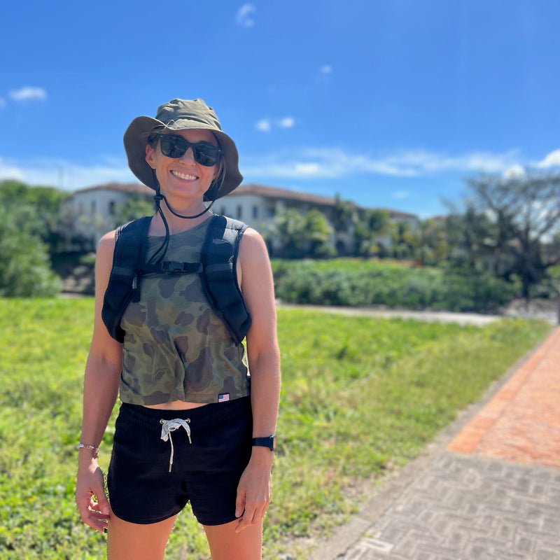 Smiling woman outdoors on a sunny day, wearing sunglasses and the Boonie Hat - Slick - ToughDry, stands on a path with greenery behind her, enjoying the comfort of its durable ToughDry fabric.