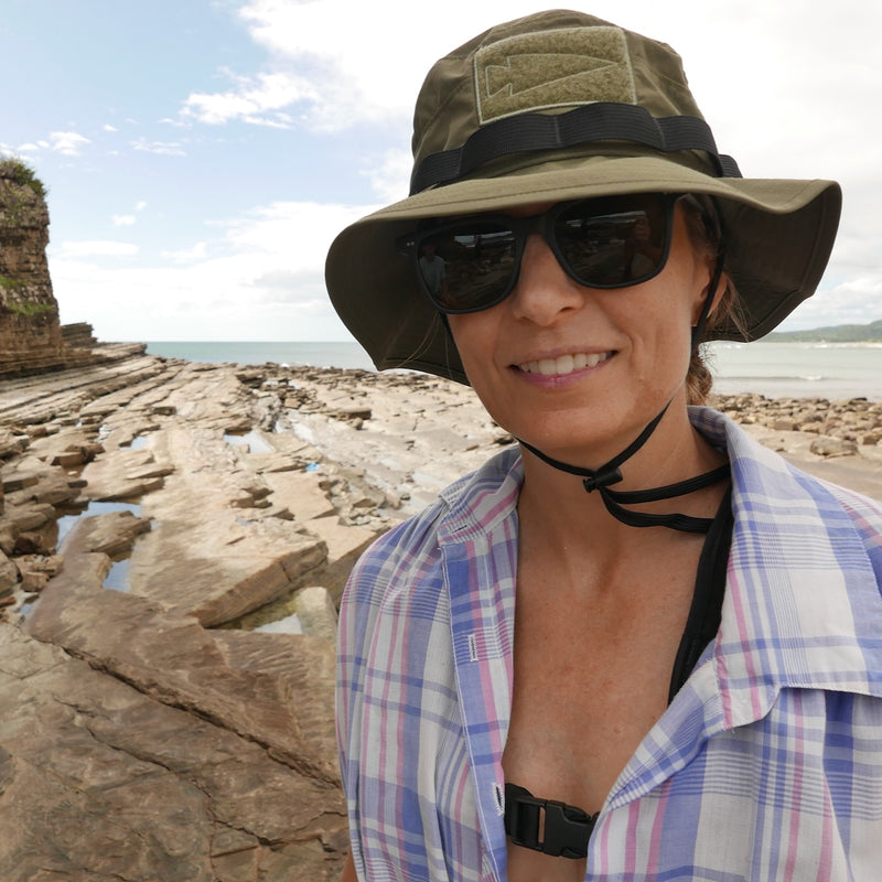 A woman in sunglasses smiles on a rocky beach with blue sky and clouds, wearing the ToughDry Tactical Boonie Hat.