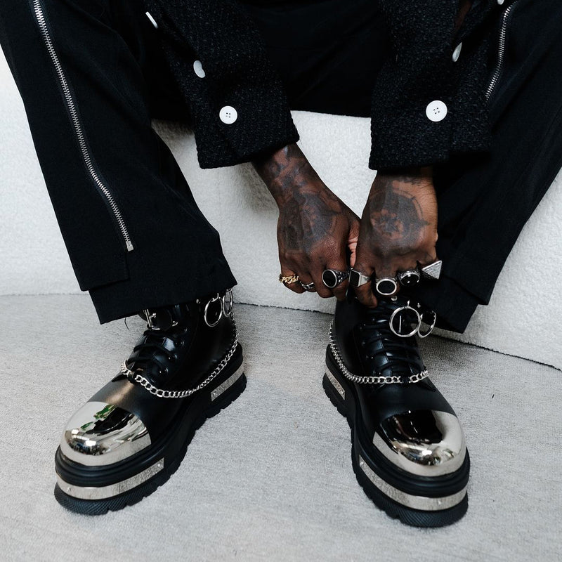 A man wearing high-top boots with chain details on a gradient background