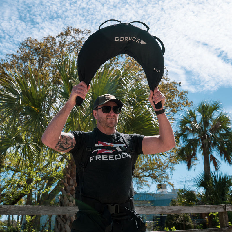 A man outdoors lifts a Bulgarian Sandbag with reinforced handles overhead, wearing sunglasses and a shirt that reads "Freedom.