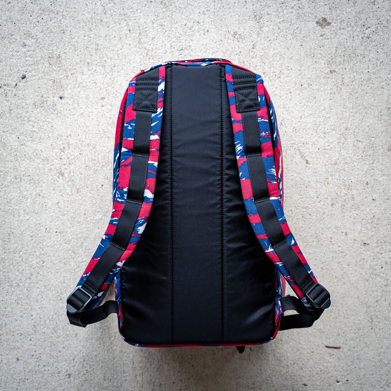 The Bullet Ruck Classic - Ballistic Nylon Cordura is a streamlined 15L daypack with blue, red, and white patterned straps and back, shown laid flat on a light concrete surface.