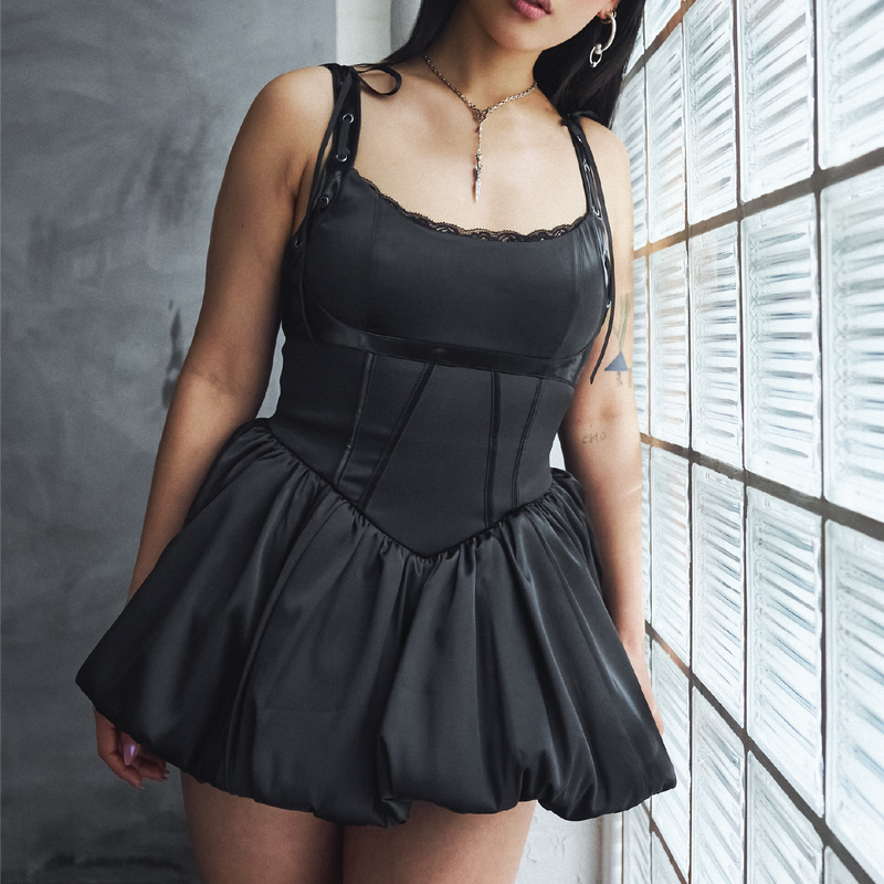 Woman wearing a black corset dress standing against a window with blinds.