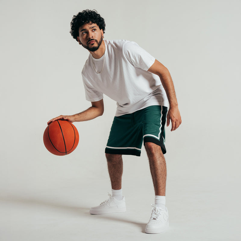 Men's green basketball shorts with black hem and white piping, worn mid-step with ball