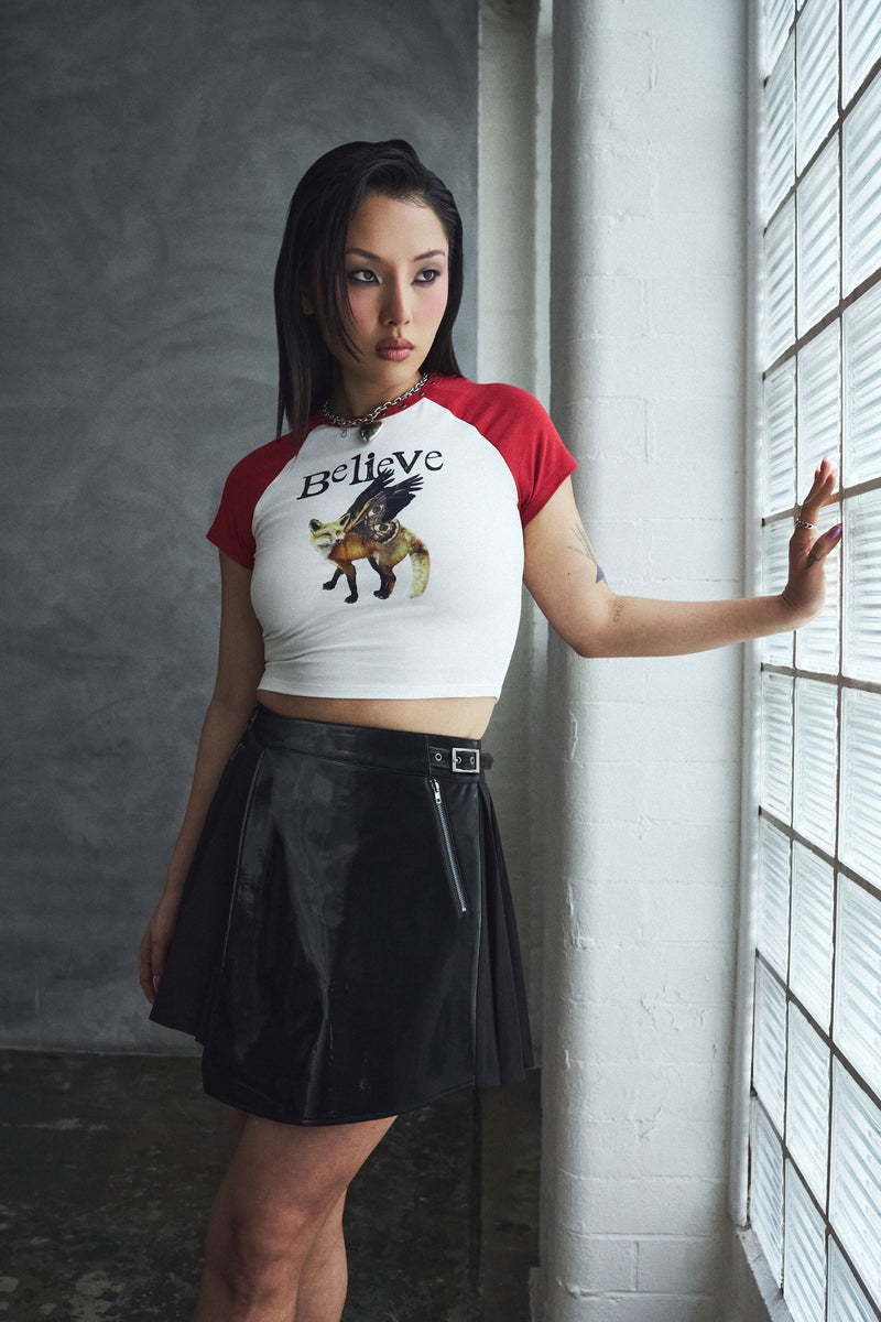 Woman wearing a white and red crop top with graphic design and black skirt, standing against a gray wall.