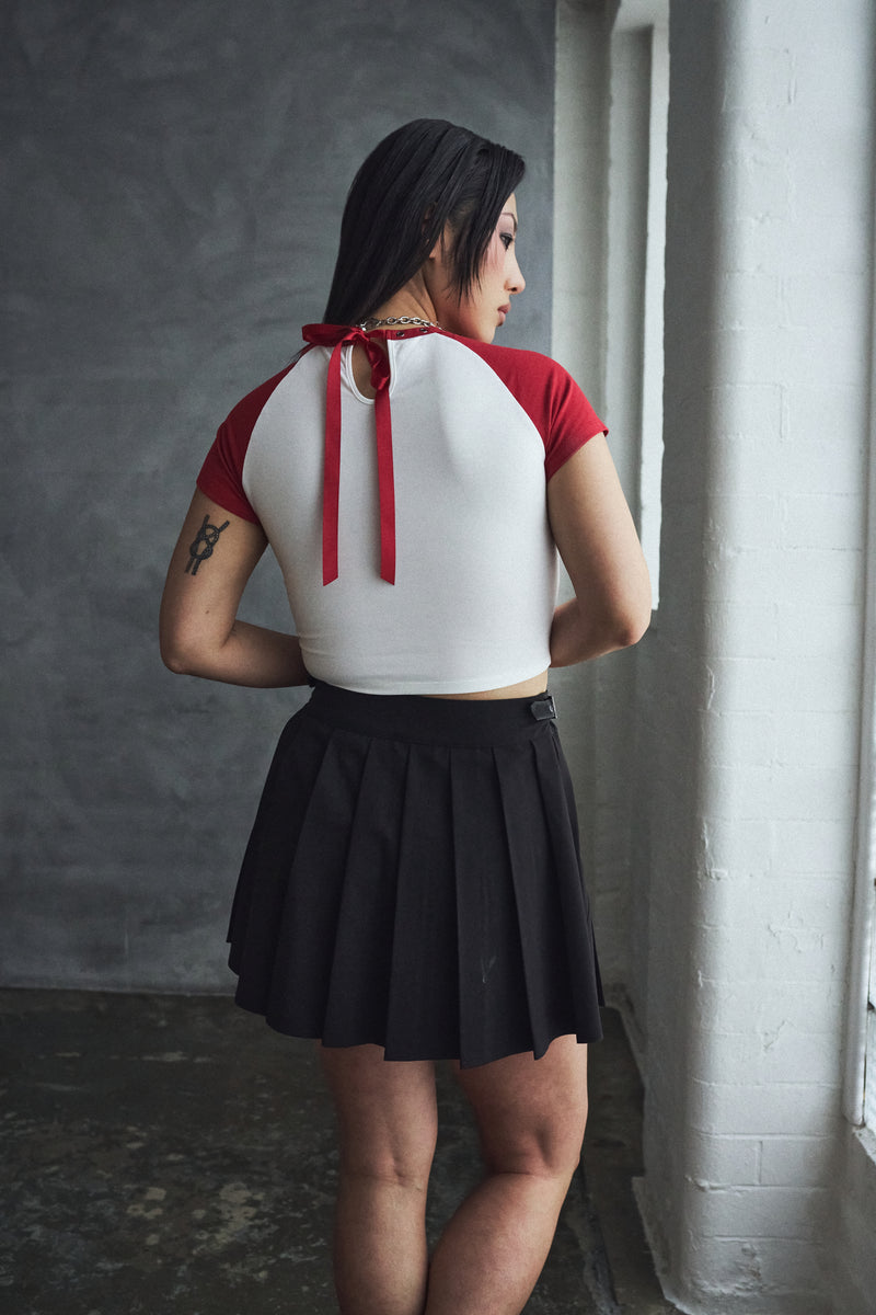 Woman wearing a white top with red accents and a black pleated skirt in a room with gray walls.