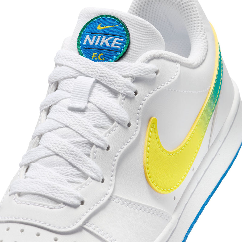 Close-up of youth white sneakers with Nike F.C. patch, yellow swoosh, green trim, laces