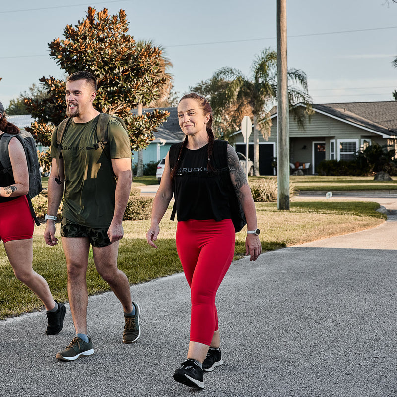 Three individuals walk down a suburban street, backpacks in tow, dressed in athletic gear. The women wear GORUCK Women's Cropped Training Leggings made from ToughFlex fabric, providing squat-proof comfort for their outdoor adventure.