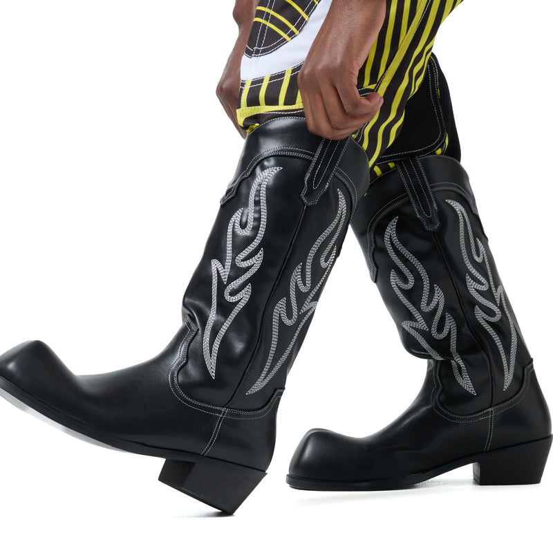 Black cowboy boots with white embroidery worn by a person in a yellow and black outfit.
