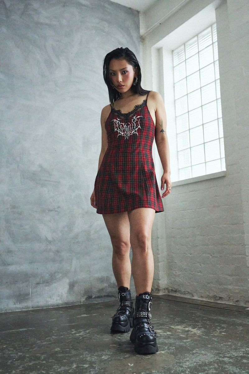 Woman wearing a red plaid dress with a black and white design, standing in a room with concrete walls and a window.
