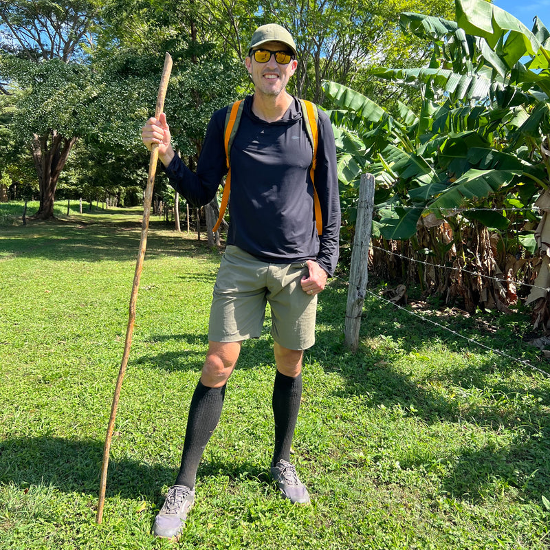 Man in outdoor gear stands on grass with a walking stick, wearing sunglasses, a backpack, and the Performance Running Hat - Italian ToughMesh made from premium Italian fabric.
