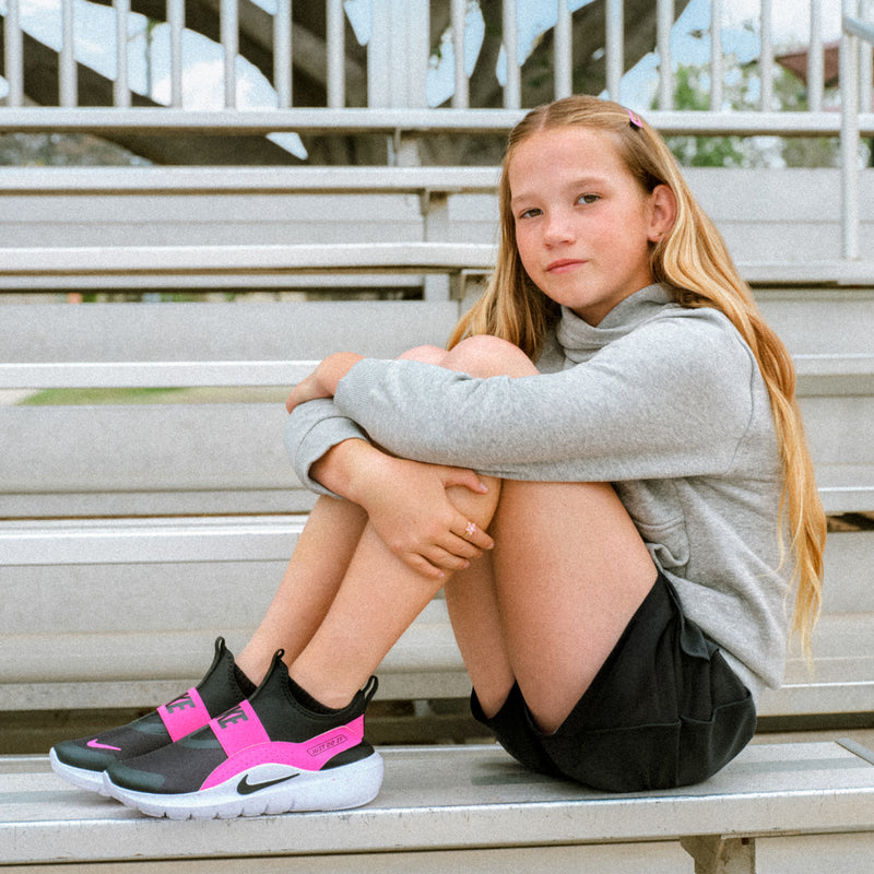 Kids slip-on running shoes on bench: black uppers, laser fuchsia straps, white soles.