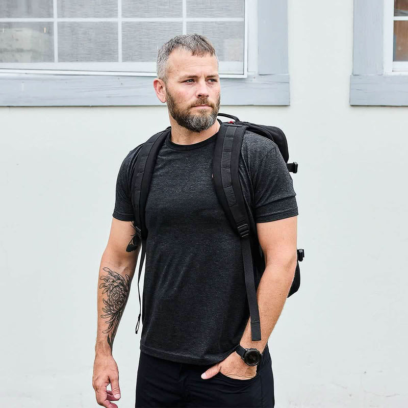 A bearded man in a black t-shirt stands outdoors near a white wall with windows, wearing the GR3 45L travel backpack.