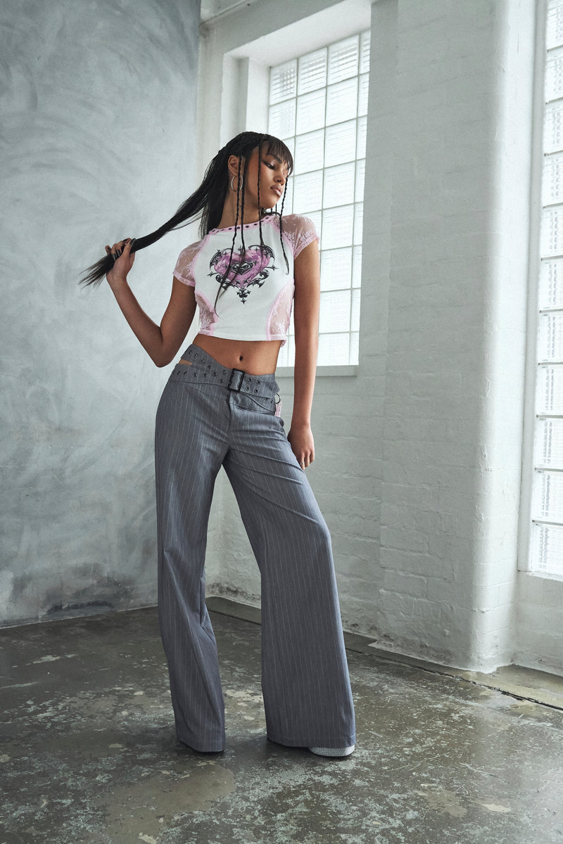 Woman in a white crop top with floral design and gray wide-leg pants standing in a room with large windows.