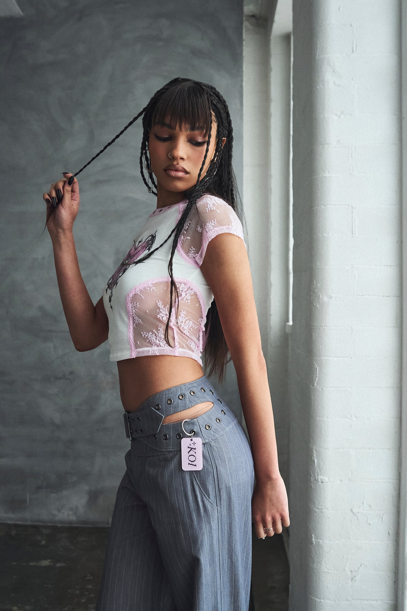 Woman wearing a white lace top and gray pants with a visible brand tag, holding a black hair tie.