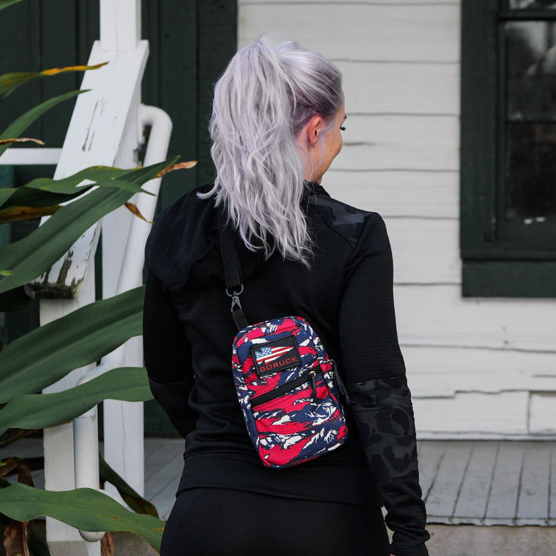 A woman with long, silver hair stands outside a white house, wearing the GRenade Ruck - American Tiger Stripe cross-body bag.