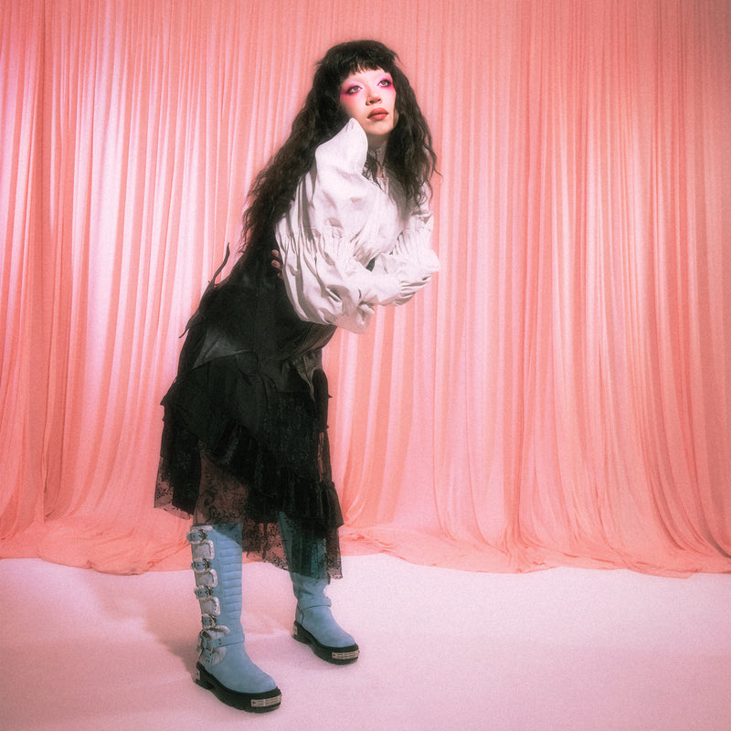 Jazmin Bean wearing a white blouse, black skirt, and blue boots against a pink curtain background