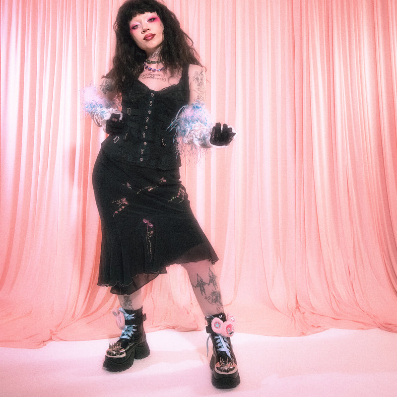 Person in gothic attire with black dress, boots, and gloves against a pink curtain background