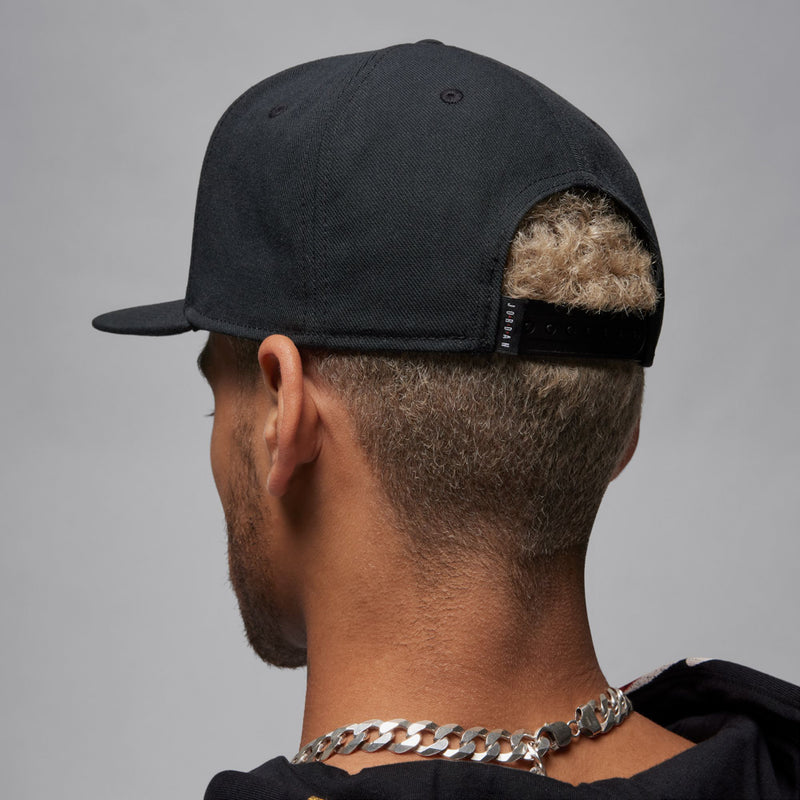 Black snapback cap back view with adjustable strap detail, worn by model.