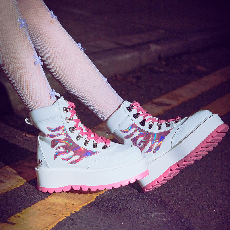 Close-up of pink and white platform boots with a holographic flame pattern and pink laces on a person's feet, set against an urban background.