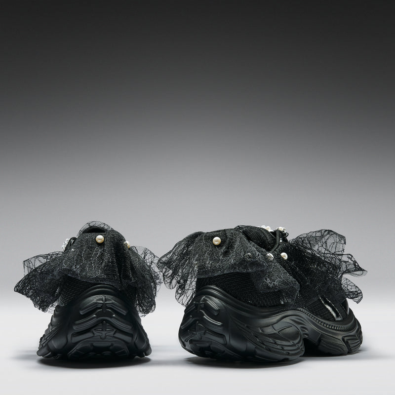 Black sneakers with sheer ruffled details and pearls on a gray background