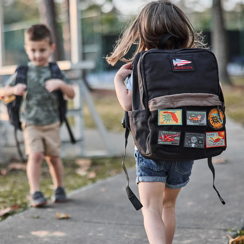 Child wearing black GORUCK backpack with colorful patches, outdoors on a sidewalk