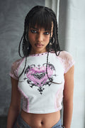 Woman wearing a white crop top with a heart design against a gray wall.