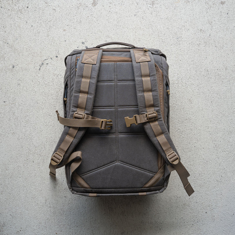 The M26 PREORDER // Q4 2026 ETA backpack, featuring padded straps, buckles, and a bombproof laptop compartment, is displayed flat on a textured light-colored surface.