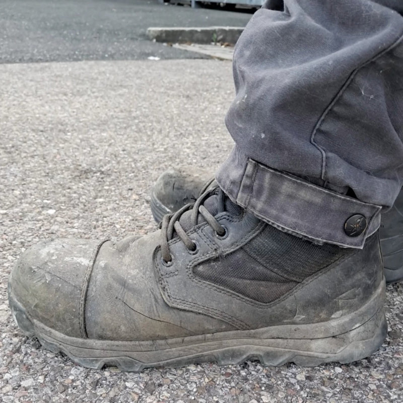 A person wearing MACV-2 Safety Boot - 8" High Top with composite toe protection and grey pants stands on a rough, gravelly pavement.