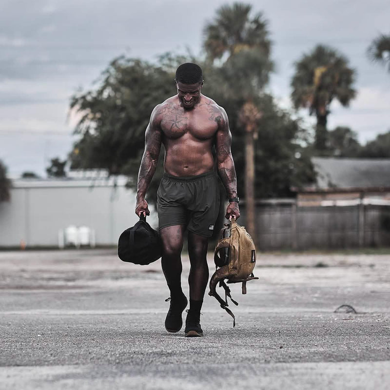 Athletic man rucking outdoors with GORUCK sandbag and backpack, palm trees in background.