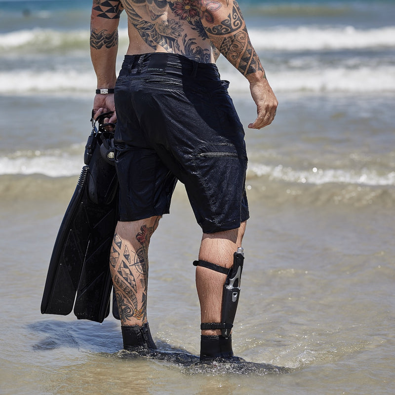 A tattooed man with a prosthetic leg, wearing Men’s Challenge Shorts - Lightweight ToughDry®, holds swim fins while standing in shallow ocean water facing the waves.