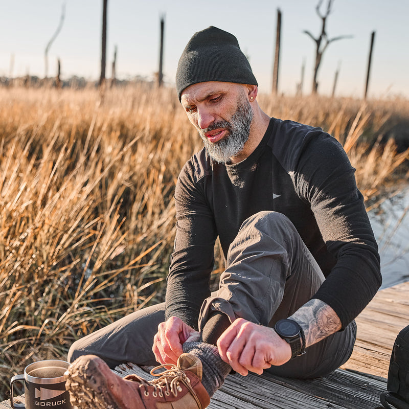 A man sits outdoors on a wooden path, adjusting his sock beside dry grass and a mug, dressed in the cozy Men’s Commando Long Sleeve - Merino Wool.