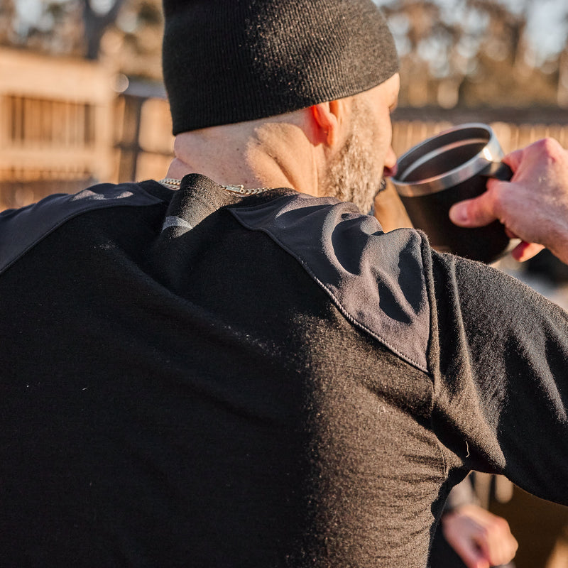 A man wearing the Men’s Commando Long Sleeve - Merino Wool and a beanie drinks from a mug outdoors, facing away from the camera.