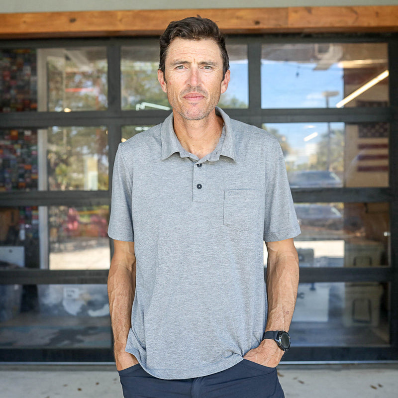 A man stands outside in front of glass doors, wearing the Men's Polo with Pocket - Tri-BlendX, hands in pockets, highlighting the style and comfort of its premium tri-blend fabric.