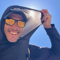 A smiling man in sunglasses and a hat pulls the hood of the Men's Sun Hoodie - ToughMesh over his head, enjoying the sun with UPF 30+ protection beneath a bright blue sky.