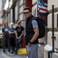 Man wearing GORUCK backpack and sunglasses standing on city street, American flag in background