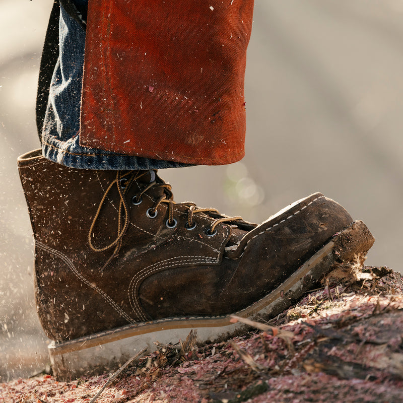 Men's dark brown lace-up work boot in muddy conditions, moc toe and tan sole visible
