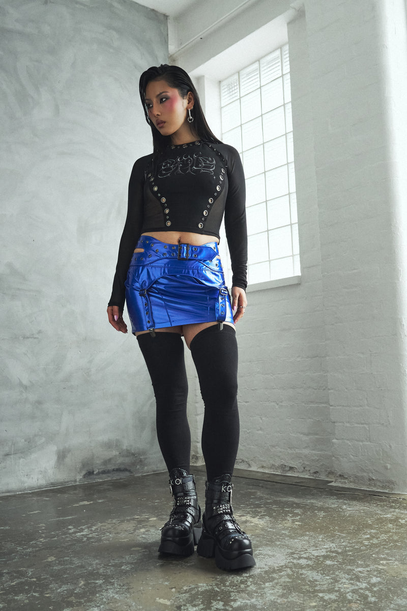 Person wearing a black long-sleeve top with button details and a blue metallic skirt in a room with a window.