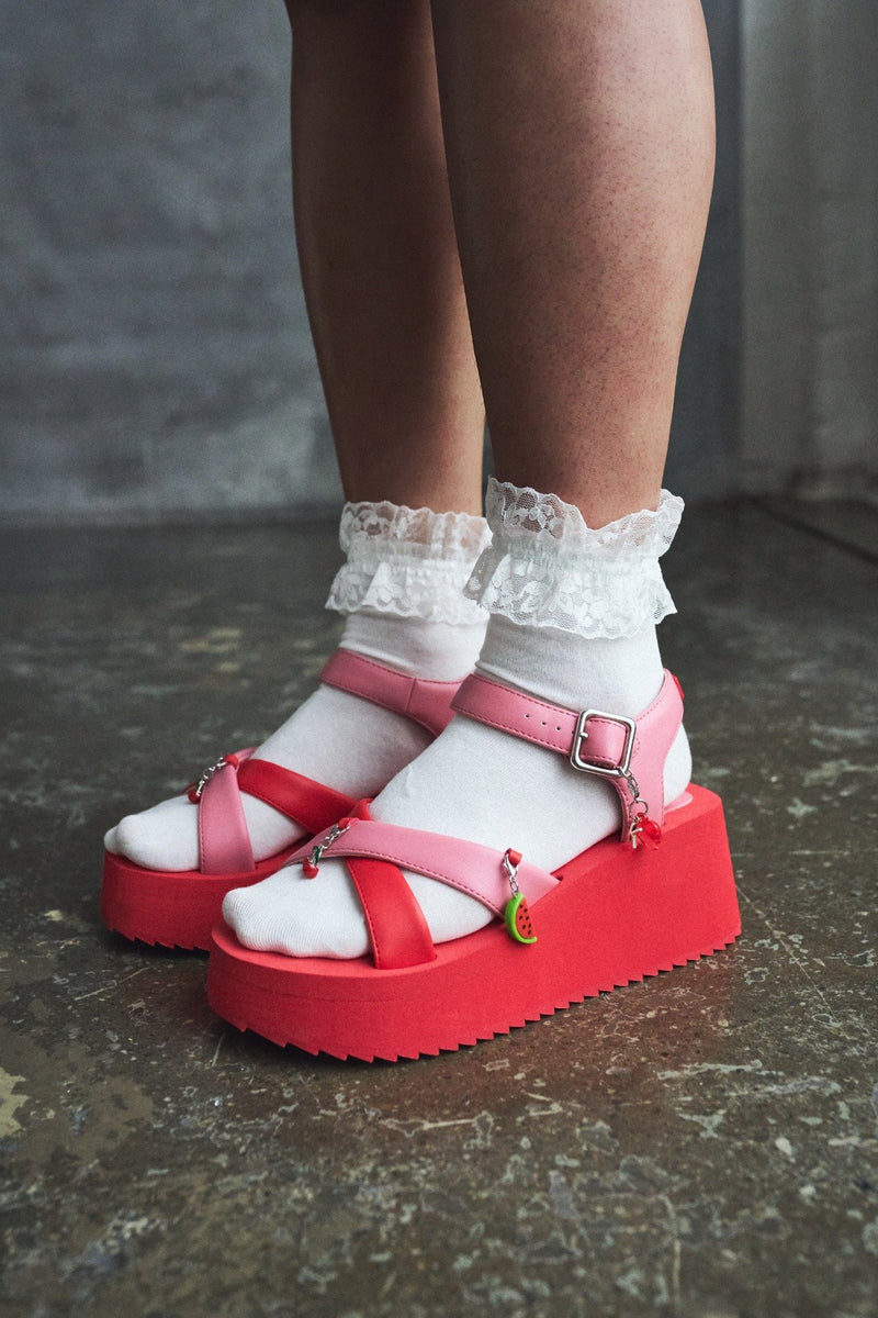 Red platform sandals with white socks and ruffled leg warmers on a dark floor.