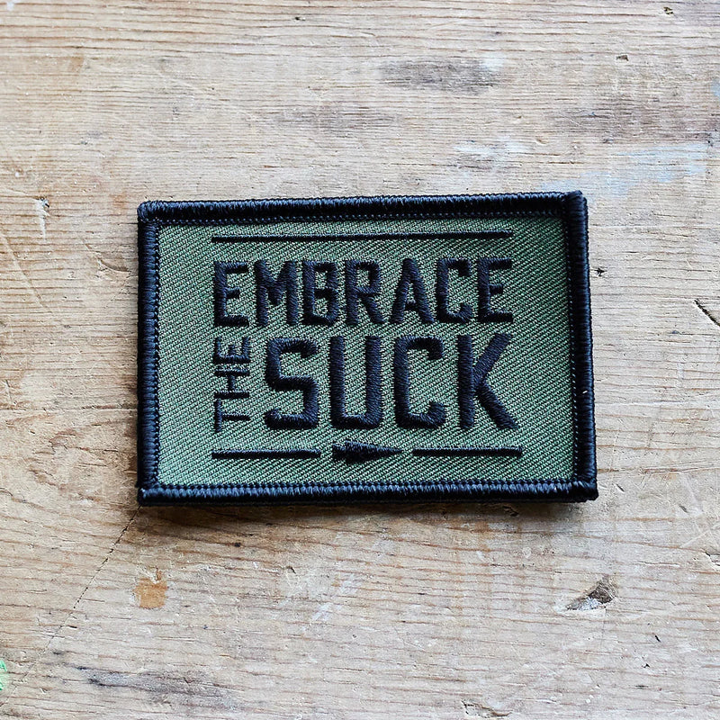 GORUCK green embroidered morale patch with 'Embrace the Suck' text on wood surface