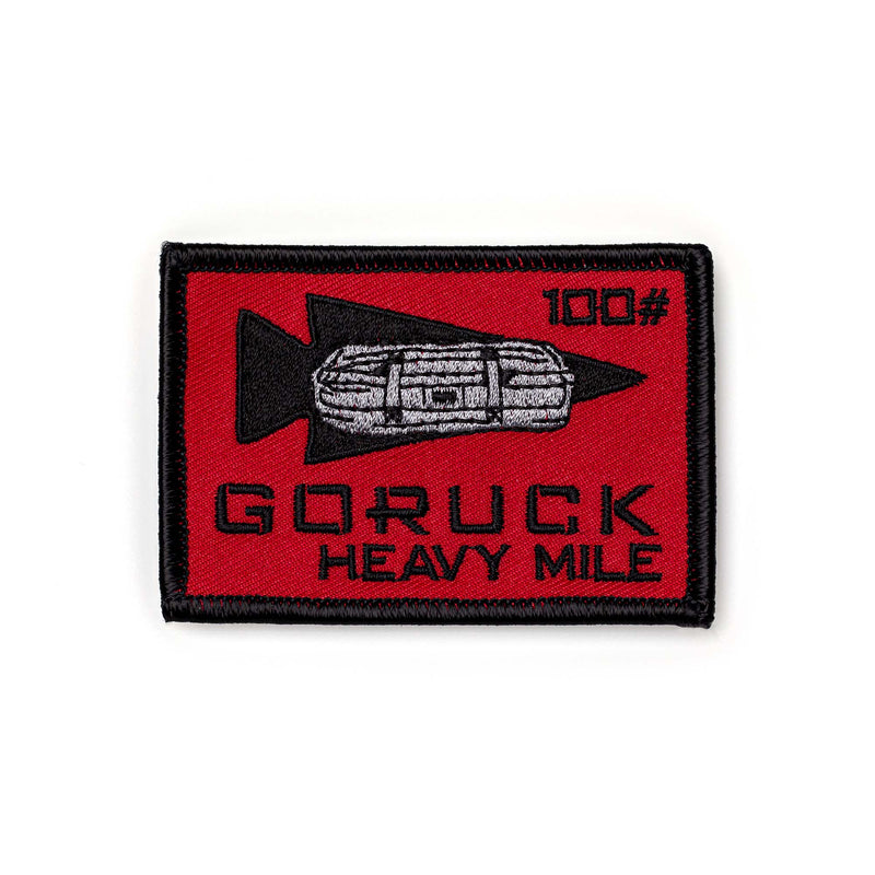 The Patch - HEAVY MILE features a red rectangle with a black arrow, sandbag, "GORUCK HEAVY MILE," and "100" text—ideal for commemorating your accomplishment in the challenging Heavy Mile weight carry event.