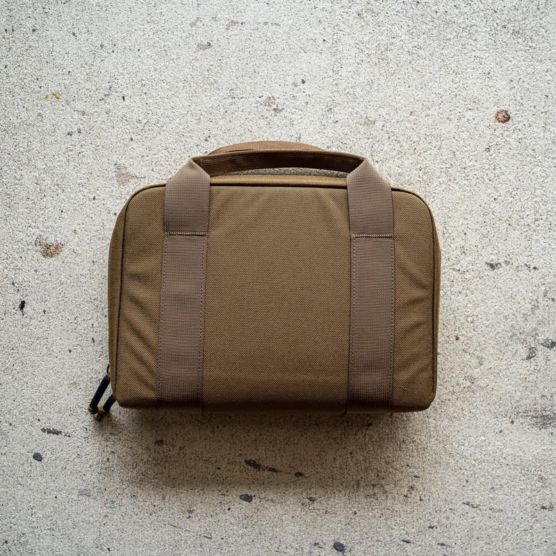 The Pistol Rug, a brown soft-sided modular pistol case with two handles, rests on a light textured concrete surface.